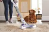 A person is vacuuming a rug with a cordless vacuum cleaner, while a golden retriever dog lies on the floor nearby. Podgląd zdjęcia produktu