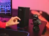 A hand is adjusting the volume knob on a black speaker next to a computer monitor and a small bust. Produktbild-Vorschau
