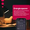 A desk lamp illuminates a sheep-shaped lamp and an open book, with a smartphone displaying a smart home app in the foreground. Produktbild-Vorschau
