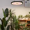 A ring-shaped LED grow light hangs above a collection of indoor plants on a wooden shelf. Náhľad obrázku produktu