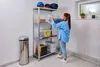 A woman is organizing items on a metal shelving unit in what appears to be a garage or storage area. A trash can is to the left of the shelving unit. Produktbild-Vorschau