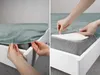 A split image showing hands putting a fitted sheet onto a mattress on a white bed frame. Produktbild-Vorschau