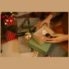 A person's hands are opening a small gift box filled with straw and three small, round items, placed on top of a larger green gift box. Several other wrapped gifts and a portion of a Christmas tree with lights are visible in the background. Podgląd zdjęcia produktu
