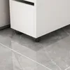 Close-up of the bottom of a white cabinet with black wheels on a gray tiled floor. Produktbild-Vorschau
