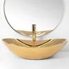 A close-up studio shot of a gold vessel sink with a gold faucet in front of a circular mirror. Náhled obrázku produktu