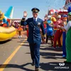 A man in a pilot costume waves to the crowd during a parade, with other costumed people and a float in the background. Anteprima dell’immagine prodotto