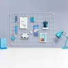 A white metal grid wall organizer with various items attached, including photos, papers, pens, and a desk lamp. Náhľad obrázku produktu