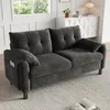 A dark gray chenille sofa is shown in a well-lit living room setting, placed on a light gray rug. A window with greenery visible outside is in the background, along with a white cabinet and a table lamp. Produktbild-Vorschau