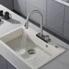 A kitchen sink with a faucet running water into it. The sink is made of a light-colored composite material and is set into a countertop. The faucet is modern and has a pull-down sprayer. The background shows kitchen cabinetry. Podgląd zdjęcia produktu