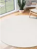 A round, white rug is placed on a wooden floor in a room with a large window overlooking a garden. A potted plant and a chair are also visible. Produktbild-Vorschau