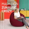 A woman sits on a large red beanbag chair in a colorful room, writing in a notebook. Produktbild-Vorschau