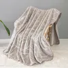 A beige blanket with a white pattern is draped over a chair, with a plant in the background and a fluffy rug in the foreground. Podgląd zdjęcia produktu