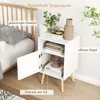 A white nightstand with an open door, placed next to a bed in a bedroom setting. The nightstand has shelves and is decorated with books and a vase. Náhled obrázku produktu