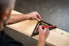 A person's hands are using a black triangular ruler and a red pencil to mark lines on a wooden plank. Voir les images du produit