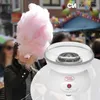 A person is holding a large piece of pink cotton candy, with a white cotton candy maker machine in the foreground. The background shows a blurred festive scene. Podgląd zdjęcia produktu