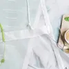 Close-up of a green embroidered sheer curtain with white trim and ties, with a small cutting board and food item in the background. Produktbild-Vorschau