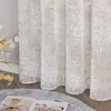 A close-up view of a floral patterned sheer curtain hanging in a room, with a book and a cup of tea on a rug in the foreground. Produktbild-Vorschau