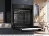 A modern kitchen scene featuring a built-in oven with its door open, showcasing the interior. The background includes wooden wall paneling and dining furniture. Produktbild-Vorschau
