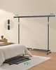 A black garment rack is shown assembled in a bedroom next to a bed. Another garment rack is shown disassembled on the floor. Náhled obrázku produktu