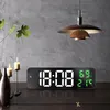 A digital alarm clock is displayed on a wooden table in a room with a textured gray wall and a pendant lamp. Voir les images du produit