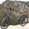 An electric bicycle is parked on a rocky shore next to a body of water, with a rocky cliff in the background. Produktbild-Vorschau