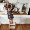 A young girl stands on a wooden step stool in a kitchen, reaching for something on a shelf. Náhľad obrázku produktu