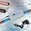 A water-themed background with water droplets, featuring a water conditioner device attached to copper pipes, along with various types of pipes and a power adapter. Voir les images du produit