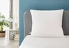 A white pillow is placed on a bed with a grey headboard and a blue wall behind it. To the left, a nightstand with a plant and books is visible. Produktbild-Vorschau