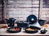 A collection of black cookware, including a tajine, Dutch oven, and fondue set, arranged on a rustic wooden table against a wooden plank background. Anteprima dell’immagine prodotto