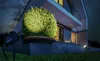 A modern house exterior at night, with garden lights illuminating a large bush and the building's facade. Náhled obrázku produktu