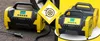 Two portable air compressors are shown, one in the foreground and one slightly behind it. A yellow diamond-shaped graphic with a tire icon is placed between them. The background is split between a dark gray asphalt-like texture and a bright yellow area. Produktbild-Vorschau