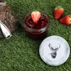 A jar of strawberry jam with a strawberry on top, placed on grass next to a piece of wood and a spoon. A lid with a deer illustration is also visible. Podgląd zdjęcia produktu