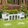 A wooden rabbit hutch with an attached outdoor run is set up on a grassy lawn in front of a wooden house. Produktbild-Vorschau