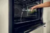 A hand is pouring liquid from a bottle into the bottom of an open oven. Produktbild-Vorschau