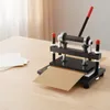 A manual die-cutting machine is shown on a wooden table, with a stack of paper to the left and a piece of brown cardstock being cut. Náhled obrázku produktu