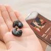 A hand holding two black wireless earbuds with a smartphone in the background displaying music. Produktbild-Vorschau