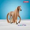 A toy horse with a long mane and tail, standing on a white surface with a light blue background. Several small grooming accessories are scattered in front of the horse. Voir les images du produit