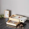 A wooden jewelry box with drawers and compartments, displaying rings and a watch, placed on a marble countertop next to a framed picture and some greenery. Voir les images du produit
