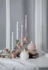 A decorative arrangement of candles and artificial roses on a white surface, possibly a windowsill, with a blurred background of white window frames and gray curtains. Produktbild-Vorschau
