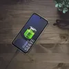 A smartphone charging on a wooden table with a plant and glasses in the background. Produktbild-Vorschau