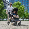 A man is pushing a stroller with a baby in it on a cobblestone street in front of a modern building. Náhled obrázku produktu