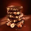 A stack of chocolate bars with hazelnuts, with loose hazelnuts scattered around the base. Produktbild-Vorschau