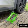 A green scissor jack and a tire iron are placed on a paved surface next to a car's tire. Produktbild-Vorschau