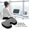 A woman works at a desk in an office with a large window overlooking a city. A seat cushion is shown in the foreground. Produktbild-Vorschau