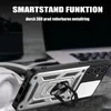 A close-up shot of a rugged smartphone case with a ring holder, placed on a textured surface with a blurred background of a laptop and headphones. Produktbild-Vorschau