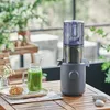 A Hurom juicer is set up on a wooden table outdoors, next to a glass of green juice and a pastry. Produktbild-Vorschau
