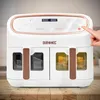 A white and rose gold dual-chamber air fryer sits on a wooden table, with a hand pressing a button on the control panel. Produktbild-Vorschau