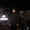A man in riding gear stands next to a horse at night. Voir les images du produit