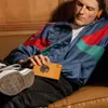 A person wearing headphones and a colorful jacket is sitting on a couch, holding a cassette tape. Náhled obrázku produktu
