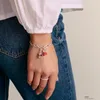 A person's hand and wrist wearing a charm bracelet, with a white shirt and blue jeans visible. Podgląd zdjęcia produktu
